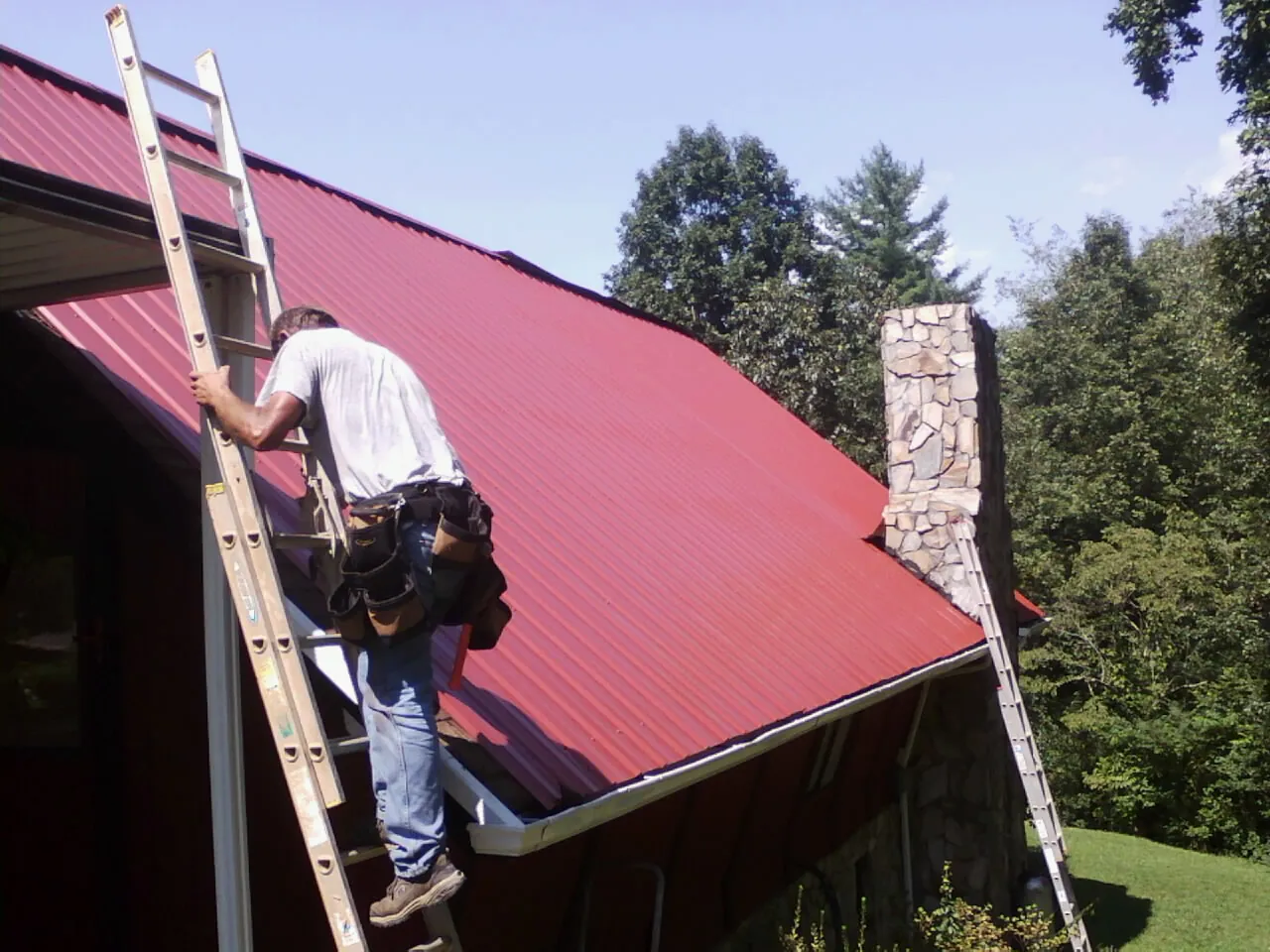 Roofer on a ladder beside a red metal roof and stone chimney — Soffit & Fascia Repair by Roofing Brothers in Abell